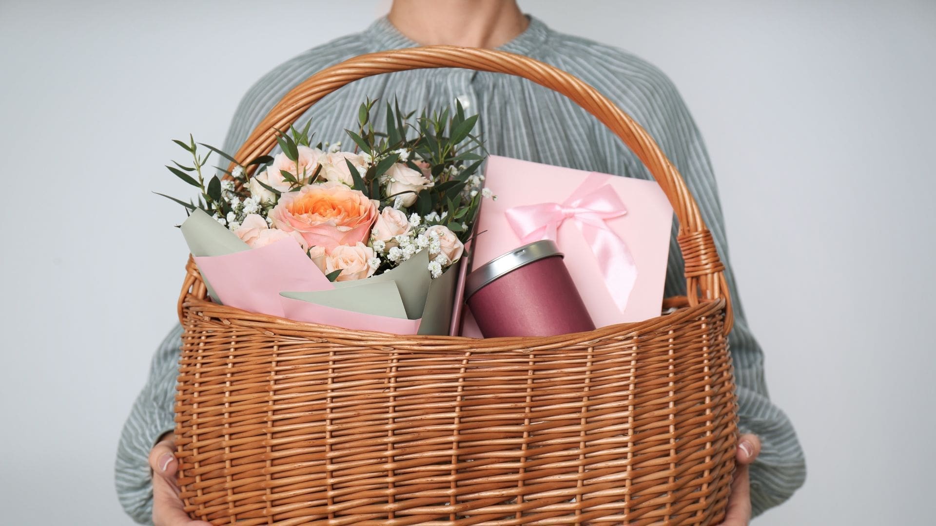 Woman Holding Wicker Basket with Different Gifts on Grey Background, Closeup